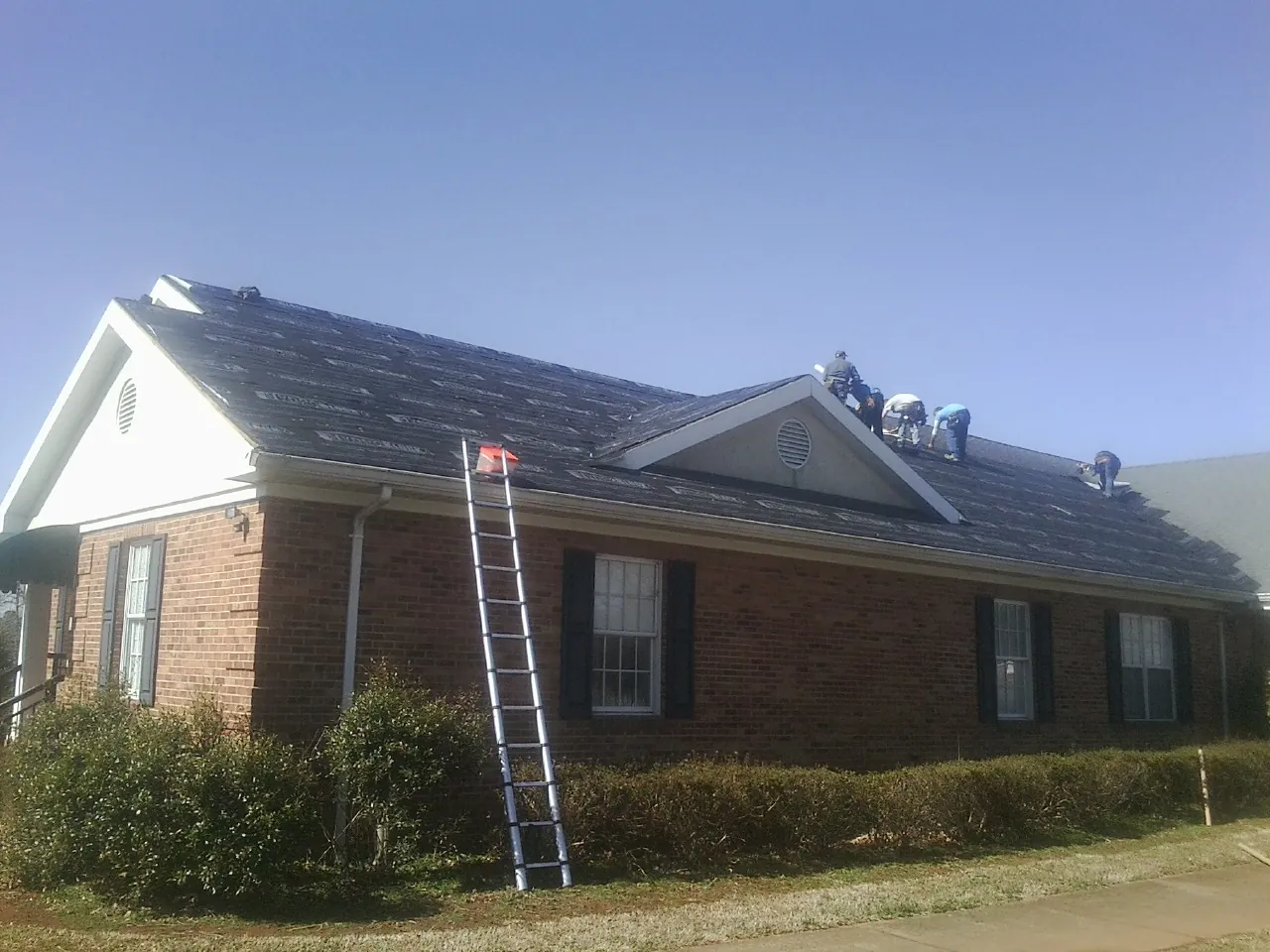 Crew tearing shingles off a brick ranch roof with a ladder set — Metal Roof Repair work by our crew in Durhamville