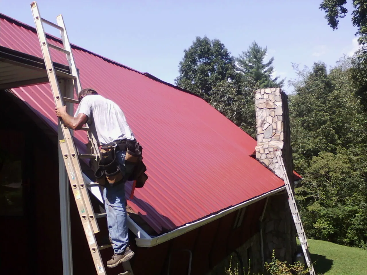 Roofer on a ladder beside a red metal roof and stone chimney — Soffit & Fascia Repair by Roofing Brothers in Durhamville